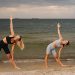 Two women exercising at a beach, performing yoga poses with the ocean in the background during sunset.
